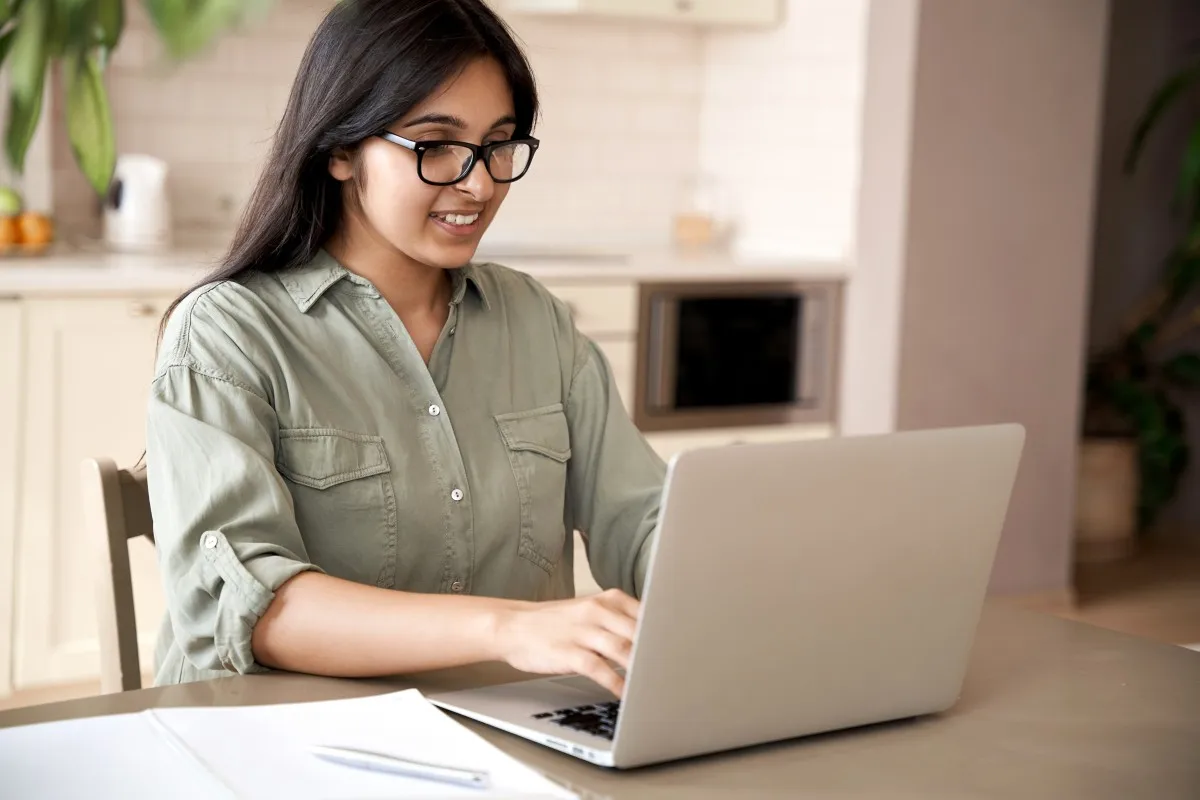 A woman smiling as she types on a laptop at a kitchen table